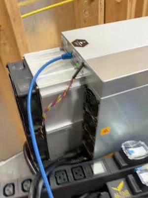 The cover image shows a close-up view of cryptocurrency mining hardware. The scene is dominated by the metallic, angular form of an Antminer miner, with visible fans and circuit details, suggesting a focus on digital finance. A blue Ethernet cable is prominently connected, highlighting the active setup. The equipment is set up indoors.
