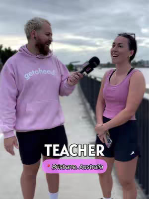 A man in a pink hoodie interviews a woman on a bridge in Brisbane, Australia. Text overlays read 'TEACHER' and 'Brisbane, Australia.'
