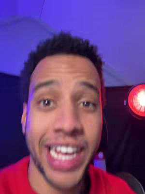 A close-up shot of a young man with short, dark, curly hair and a red shirt. He is smiling widely, showing his teeth, and appears to be speaking. The background is dark but has blue and red lighting, with a ring light visible on the right side.