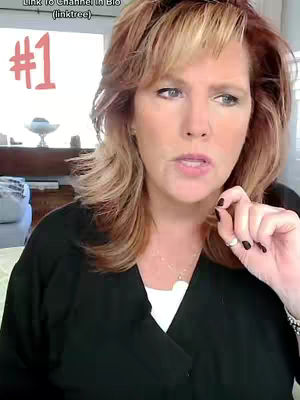 A woman with light skin and reddish-brown hair is centered in the frame. She wears a black top over a white undershirt. The background includes a window and elements of a room. In the top left corner, there is a white area with red text and below that, there's text in the top left corner. She is holding one hand slightly up near her mouth and looking towards the upper right.