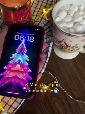 The image features a smartphone displaying a colorful Christmas tree charging animation, held by a hand. It is set on a wooden table next to a mug of marshmallows, a lit candle, and Christmas lights.