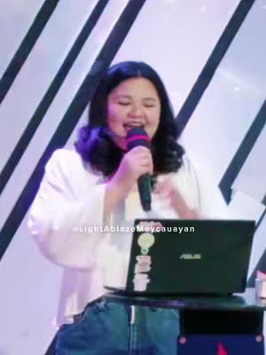 A woman with dark wavy hair, wearing a white blouse and denim jeans, speaks into a microphone, her hands gesturing expressively. She stands in front of a backdrop with white and black diagonal lines. A laptop showing an ASUS logo with stickers sits on a small table in front of her, and a microphone in her hand. The image is brightly and evenly lit, and the overall composition centers on the speaker.