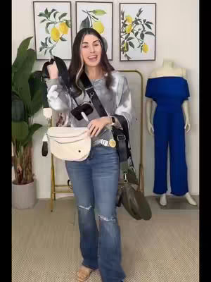 A woman stands in a well-lit room, holding a white, crescent-shaped bag. She wears a tie-dye sweater, ripped jeans, and carries other bags with long straps. Behind her are three framed lemon art pieces and a mannequin wearing a solid dark blue jumpsuit. A green houseplant in a pot and a clothes rack add depth to the scene.