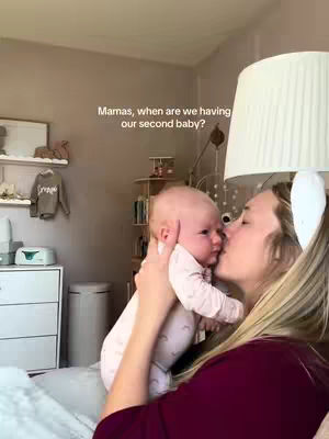 A woman holds a baby close, kissing its cheek. The baby is facing towards the camera. The woman has blonde hair and is wearing a burgundy top. The background is a room with neutral colors and a lamp.