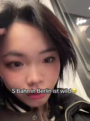 A selfie-style shot featuring a young woman inside a public transport coach, likely a train. She is the central focus, with the interior of the coach subtly visible in the background. The woman has dark hair and is looking directly at the camera with an expression that appears to be a mix of surprise and amusement. The lighting seems to come from the interior lights of the transport.