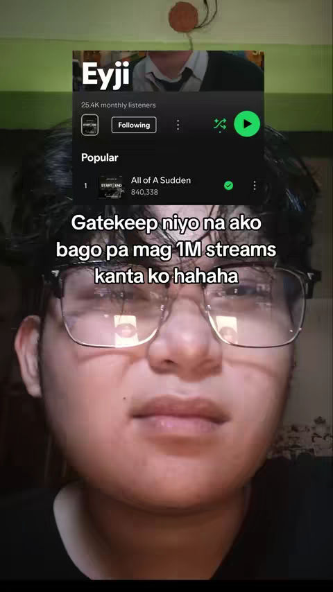 A selfie-style shot featuring a young man in glasses, framed from the chest up. He's looking towards the camera with a slight smile or neutral expression. Behind him, a Spotify interface is visible on a screen, showcasing an artist profile with the name 'Eyji,' their number of monthly listeners, and a music player displaying a song, 'All of a Sudden.' Overlaid on the image is the text "Gatekeep niyo na ako bago pa mag 1M streams kanta ko hahaha" in a white font. The background has a blurry, muted green and brown color scheme.
