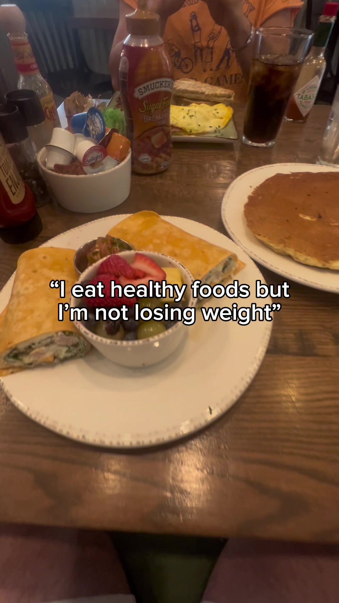 The image shows a table with various food items and condiments. In the foreground, there is a plate with a wrapped breakfast item, a small dish of fruit, and a text overlay with words related to dieting. On the right side, there is a pancake and glass. Other condiments can be seen in the background.