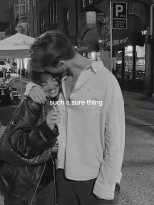A black and white photo shows a couple embracing on a city street. The man, on the right, is wearing a button-down shirt and appears to be gently touching the woman’s head. The woman is smiling broadly, wearing a dark leather jacket, and has her arm around the man.