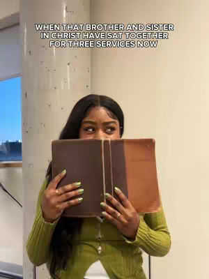 A woman with long black hair is holding a book in front of her face, obscuring most of it. She is wearing a green long-sleeve shirt and there is text at the top of the image. The background suggests a neutral, indoor setting, with a glimpse of a window on the left side.