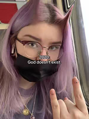 A selfie-style shot featuring a young person with a light purple hair, rimless glasses, and holding up two fingers. The background appears to be a train setting with other visible. Text overlayed on the image says, "2020: God doesn't exist."