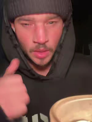 A medium shot of a young man wearing a black beanie and hoodie. He has a short beard, a nose ring, and is looking towards the side with what appears to be a slight smile. He is giving a thumbs-up gesture with his left hand. The lighting is dim.