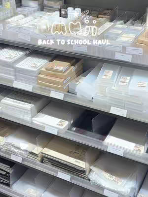 A photo of the interior of a Muji store, showcasing rows of shelves filled with various stationery and organizational products. The shelves are made of a light-colored wood, and the products appear to be neatly arranged with labels and price tags. The lighting is bright, and the overall aesthetic is clean and minimalist.