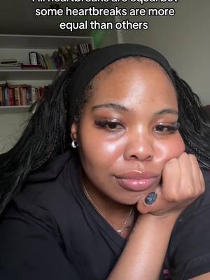 A close-up shot of a woman with dark hair and a black headband. The woman is looking at the camera with a soft, almost sad expression, and her hand is resting on her cheek. Above her head, there's text that reads: "All heartbreaks are equal but some heartbreaks are more equal than others."