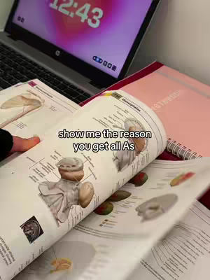 The photo shows a laptop with a brightly lit screen displaying the time, accompanied by an open textbook with medical illustrations. A pink notebook is casually placed on top. Additional open pages of the book reveal more diagrams and text. The overall composition suggests a study environment.