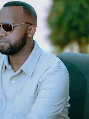 A man with short dark hair, sunglasses, and a beard is seen from the shoulder up. He's wearing a light-colored button-down shirt and appears to be outdoors, with a blurred green background.