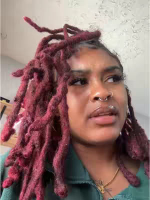A close-up shot of a young Black woman with reddish-brown locs, a nose ring, and a lip piercing. She's wearing a green top and gazing off to the side with a slightly concerned expression.