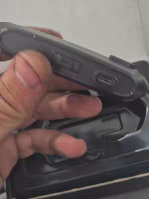 A hand holding a black device with a USB-C port, possibly a charger or a small gadget. The device sits in a black container. The background appears to be a light, neutral surface.