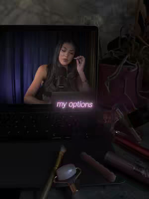 A woman sits in front of a laptop open on a desk with lipsticks, a face roller, and brush visible. A neon sign reads 'my options'. The image has a moody, dim lighting.