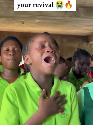 A group of people, predominantly women, are gathered indoors with one woman in the foreground. She is the focal point of the image. She has dark skin, black hair, and is wearing a green collared shirt. Her mouth is open in a cry of emotion and her eyes are closed. Her right hand is placed over her chest. The image is brightly lit with other people behind her.