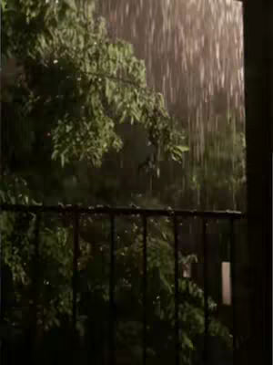 The image is a shot of a rain storm. The foreground features a black metal railing, while the background shows lush green foliage. Rain streaks diagonally across the frame, suggesting movement and weather conditions. The lighting is dim, creating a moody ambiance.