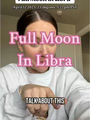 A selfie-style shot featuring a woman with fair skin and dark hair, centered against a softly lit background. Above her, text overlays state "Full Moon In Libra" and the date and time. She is pointing towards the viewer, with an added overlay "TALK ABOUT THIS". The overall composition is direct and engaging, using a neutral color palette.
