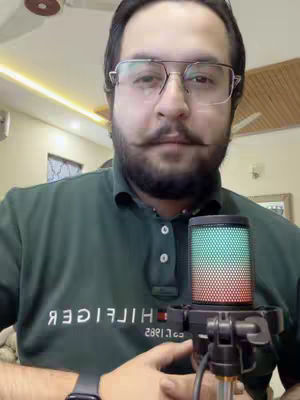 A man with glasses and a beard holds a microphone, likely in a home studio setup. The background includes a ceiling fan and light-colored walls, suggesting an indoor environment. He is wearing a green polo shirt.