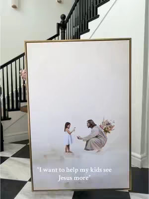 A large framed art piece is displayed against a white wall. The artwork depicts a young girl offering a flower to a figure resembling Jesus, who kneels before her. The background appears soft and light, possibly painted as a cloudy sky or heavenly setting. The frame is gold, and the art piece is placed in front of a staircase with black railing.