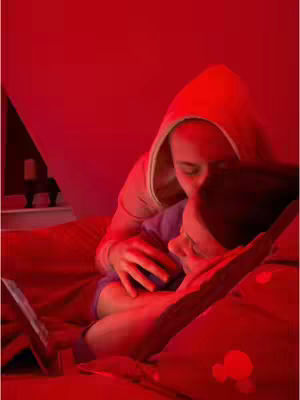 A close-up shot of two people in bed, cuddled together. The scene is bathed in a red glow, creating an intimate and warm atmosphere with a tablet in the foreground.