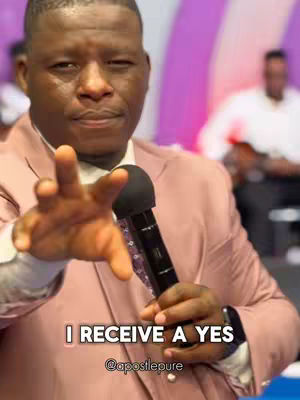 A man of African descent dressed in a pink suit extends his right arm, hand open, toward the viewer while holding a microphone in his left hand. Text overlayed in the lower portion reads 'I RECEIVE A YES' with a username below. The background is slightly blurred.