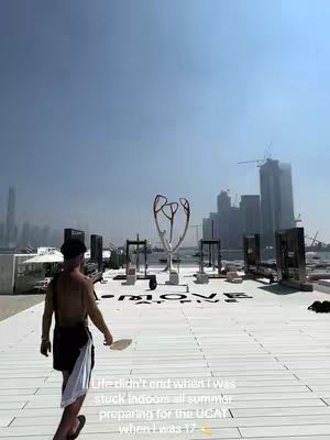 A sunny outdoor shot featuring a man walking away from the camera on a white wooden deck. Buildings and a stylized metal sculpture are in the background.
