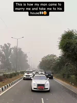 A convoy of luxury cars, including Rolls Royces, is driving down a road. The lead car is a white Rolls Royce adorned with a floral arrangement on the hood. The sky is overcast.