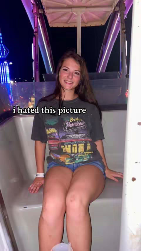 A young woman sits on a Ferris wheel, looking slightly off-camera. She wears a black graphic t-shirt, jean shorts, and a white wristband. The background shows the night sky and the illuminated structure of the Ferris wheel.