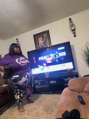 A woman in a purple t-shirt and patterned leggings is standing in front of a TV. The TV is showing a fitness game. The room has a beige carpet, a brown couch, and a remote control is on the couch.