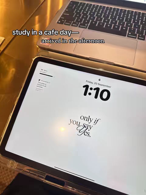 Overhead shot of an iPad and a laptop computer on a reflective surface. The iPad displays a white background with text in a stylized font. The laptop is silver with the keyboard visible and the screen lid up. The text overlay on the image reads 'study in a cafe day—arrived in the afternoon'.