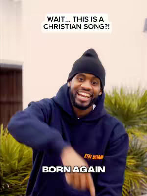A man wearing a black beanie and a blue hoodie is smiling and pointing down while standing in front of green bushes and a wall. The image contains the text overlay "WAIT... THIS IS A CHRISTIAN SONG?!" at the top and "BORN AGAIN" below the man.