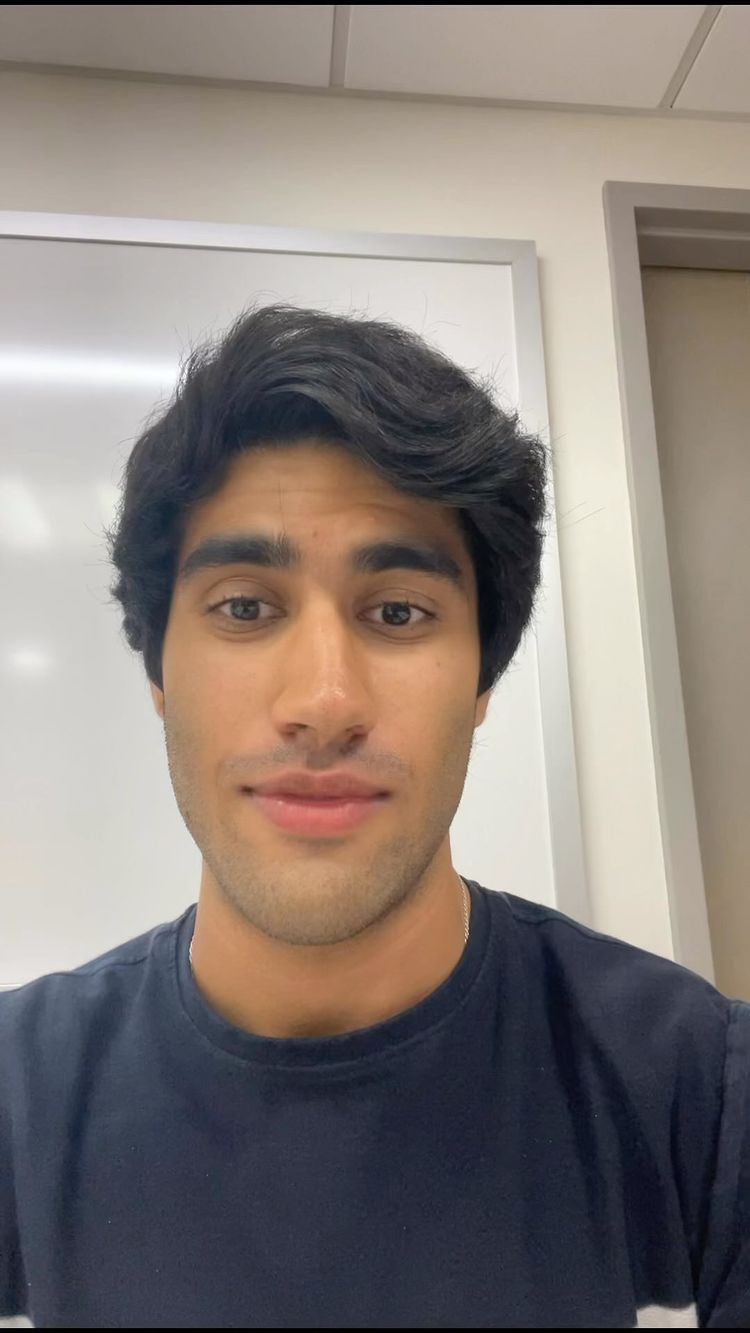 A selfie-style shot of a young man with dark hair and a dark blue t-shirt. He is centered in the frame, facing the camera directly with a neutral expression. The background is a plain, white wall or panel, and the lighting appears to be soft and even.