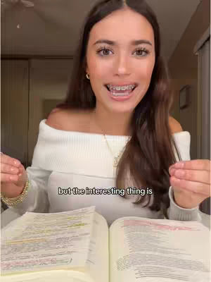 A woman with long brown hair and braces smiles widely at the camera, wearing a white off-the-shoulder sweater. She sits at a table with an open book in front of her and is holding it with her hands. The background is a blurred room setting, suggesting a comfortable indoor environment.
