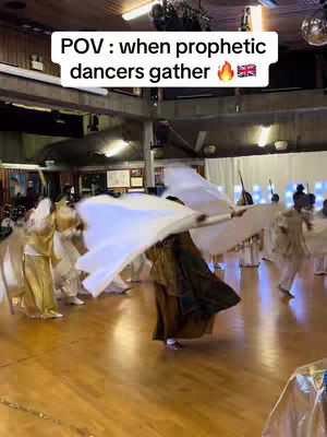 The image captures a group of people dancing in what appears to be a large hall with a wooden floor and high ceilings. The dancers are dressed in flowing white and golden-yellow attire, with some wearing white fabric that they are holding up, creating a dynamic sense of movement. The lighting suggests an indoor setting. The words "POV : when prophetic dancers gather" are in a white rectangle at the top of the image.