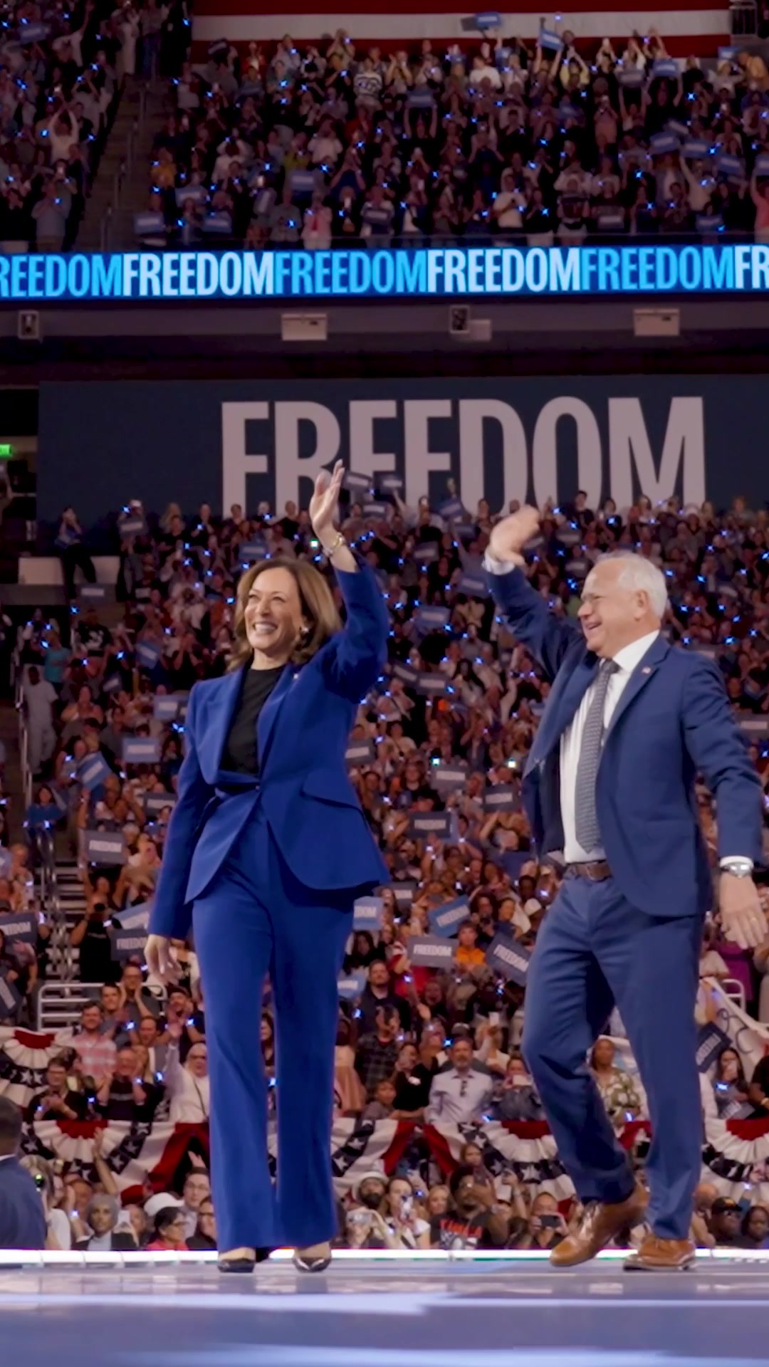 The cover image shows a crowded auditorium with a large screen reading "FREEDOM" behind a stage. Two people, a woman in a blue pantsuit and a man in a blue suit, are in the foreground, waving at the crowd. The crowd is full of people holding up lighted phones, and the overall scene suggests a political rally or celebration.