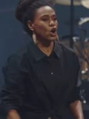 A woman speaking with an open mouth is the central focus of the image. The image has a dim lighting scheme, with a dark background that enhances the illuminated face, and the lighting comes from the top left, creating shadows on the face.
