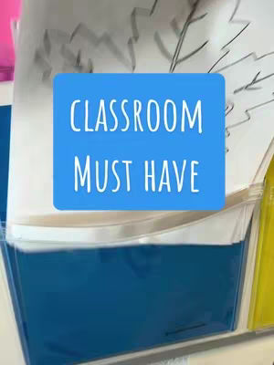 The cover image shows a close-up of a clear plastic pocket or organizer, likely affixed to a wall. Inside the pocket are several sheets of paper, one with black marker drawings on it. A blue square with white text overlays the center, reading "CLASSROOM MUST HAVE". The composition is simple, focusing on the functionality of the storage item, with a soft, out-of-focus background.