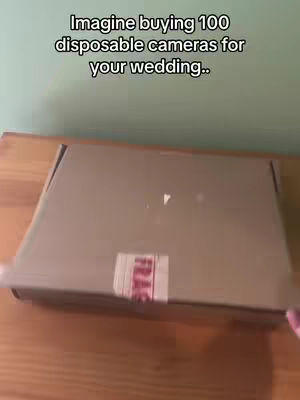 A brown cardboard box is placed on a light brown wooden surface. A section of the box's lid is lifted, revealing a red and white "FRAGILE" sticker.