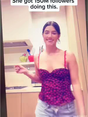 A woman is standing in a kitchen, smiling, and gesturing with one hand. The background includes kitchen cabinets, appliances, and open shelving.