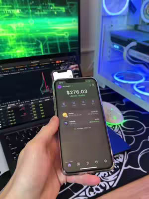 The image displays a person's hand holding a smartphone, showcasing a financial app interface with a balance of $276.03. A laptop displaying a stock market chart is slightly visible in the background, along with a desktop computer setup. The composition is well-lit, highlighting the app's user interface and emphasizing financial data and investing.