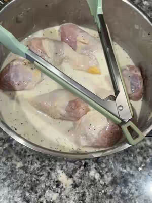 The image shows an overhead view of a stainless steel pot filled with raw chicken pieces submerged in a creamy, opaque batter. A pair of green-handled tongs rest diagonally across the pot, with one side resting in the batter. The chicken pieces have pink and tan hues, while the batter is speckled with small black dots, likely pepper. The pot sits on a gray-speckled countertop.