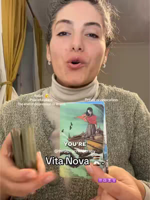A woman with fair skin and dark hair holds a tarot card and a deck of cards in front of her. The tarot card displays a drawing of a person on a boat with a scenic background, while the woman looks directly at the camera with an open mouth as if she's speaking.