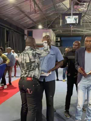 Two men are in an indoor setting with many people. One man is touching the other's forehead. The background consists of people, ceiling structures, walls, and stage lights.