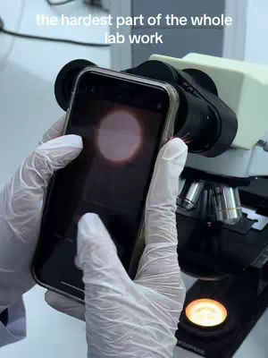 The image shows a close-up view of a person's gloved hands holding a smartphone up to the eyepiece of a microscope. The phone's screen is displaying a bright circular image, presumably what the microscope is viewing. The microscope is black and silver, and the background is a laboratory setting.