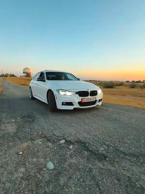 A white BMW sedan is angled slightly towards the viewer, positioned on a cracked asphalt road. The car's headlights are on, and the license plate is partially visible. The background features a gradient sky with hues of blue and orange, suggesting either sunrise or sunset with some green landscape in the background.