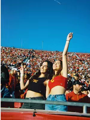 Two young women are in a packed stadium, likely at a sporting event. One is wearing a black top with text, the other a red top, and both have their arms raised in celebration. They are in front of a crowd of people, with a bright blue sky overhead, and appear to be smiling looking at the camera.
